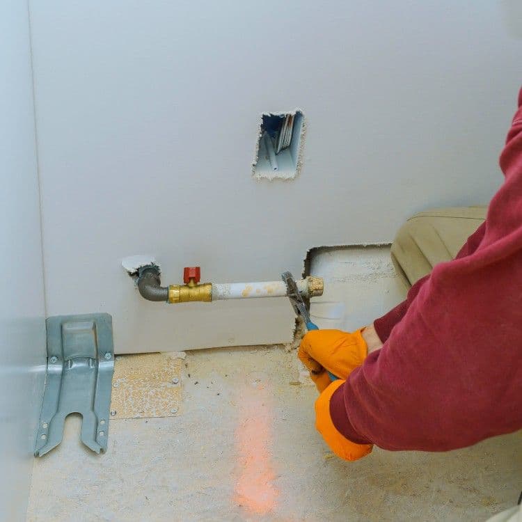 Plumber repairing a water pipe under a wall with orange gloves and tools.