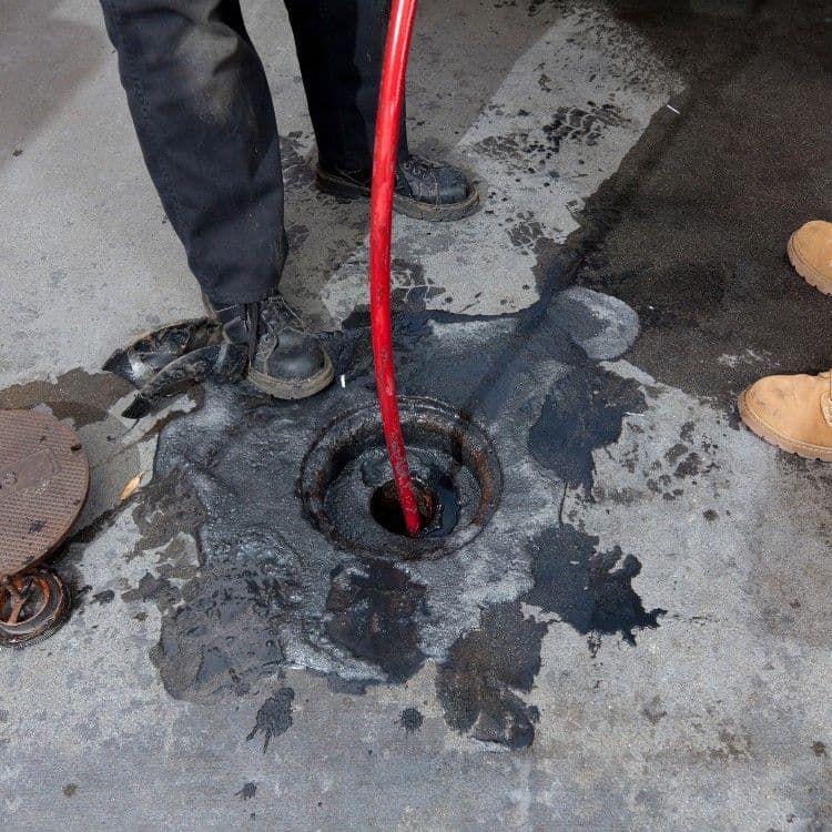 Red hose extracting liquid from a dark, stained drain on a concrete surface.