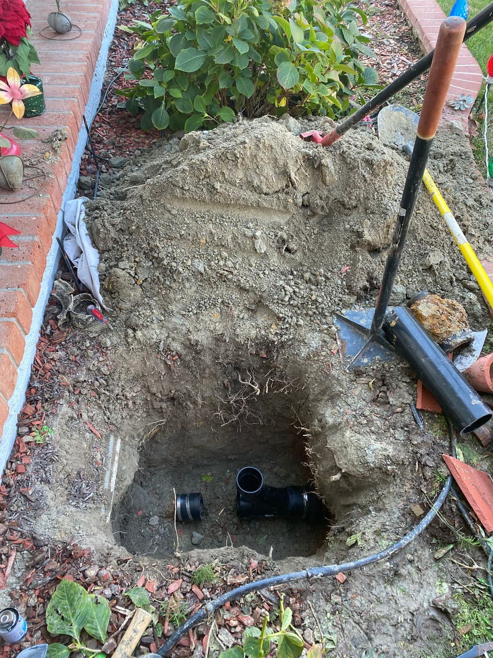 Excavated area for plumbing installation showing a black pipe and tools in a garden setting.
